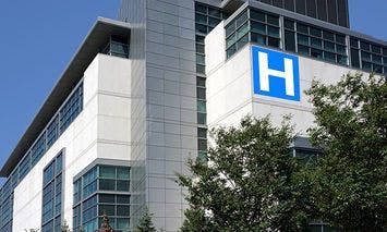 A large modern hospital building with glass windows sits against a clear blue sky, marked by a prominent blue sign with a white 
